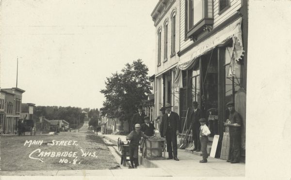 Photographic postcard view of Main Street. Several men and boys are outside of a store with H.A. Olsen printed on the awning. One boy is holding a ball. The store appears to sell hardware, machinery and ammunition, as that is what is displayed on the sidewalk. Caption reads: "Main Street, Cambridge, Wis." 