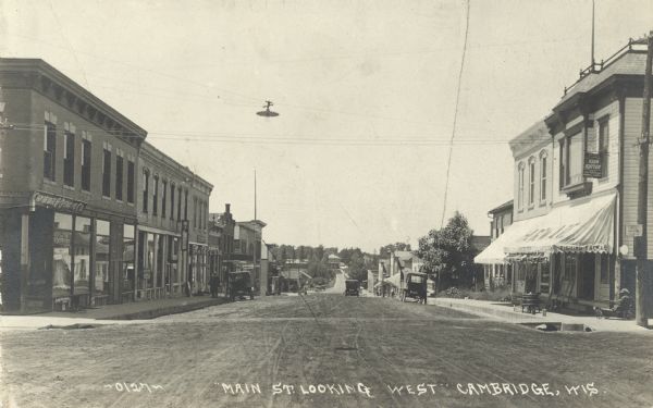 Photographic postcard of Main Street looking west. The street is unpaved and lined with many stores. Signs are advertising hardware, meat, flour and many other products. Automobiles are parked along the curb and a few pedestrians are on the sidewalks. Caption reads: "Main St. Looking West, Cambridge, Wis."