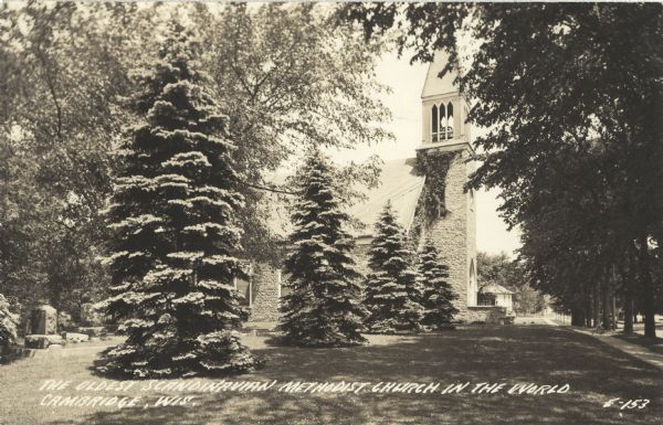 Photographic postcard of the oldest Scandinavian Methodist Church in the world. Four large pine trees are growing in the foreground and a tree-shaded side walk is on the right. On the left are some grave stones. Caption reads: "The Oldest Scandinavian Methodist Church in the World, Cambridge, Wis."