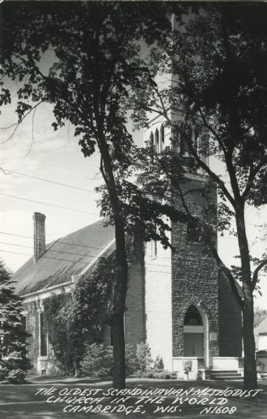 Photographic postcard of the oldest Scandinavian Methodist Church in the world. Two large trees frame the front entrance, with shrubs and trees are around the church. Caption reads: "The Oldest Scandinavian Methodist Church in the World, Cambridge, Wis."