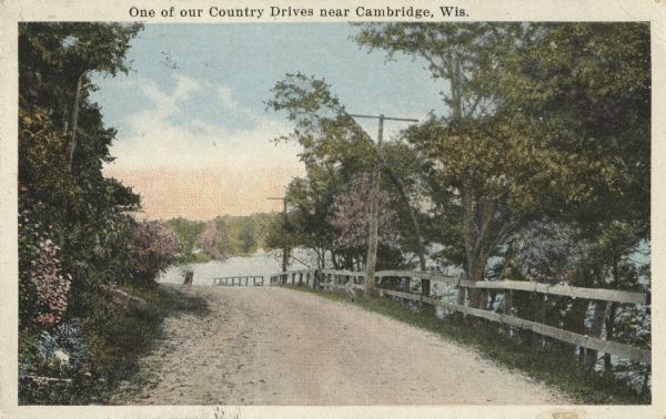 Colorized postcard of a dirt road skirting a body of water or river. A guard fence can be seen between the road and the water. Text at top reads: "One of our Country Drives near Cambridge, Wis."