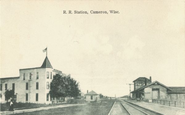 Black and white postcard of a railroad station across from the Hotel Wells. Railroad tracks run between them, disappearing into the distance. There are trees in front of the hotel and a flag is flying from the roof. A woman is standing on the sidewalk in the lower left corner. Text at top reads: "R. R. Station, Cameron, Wisc."
