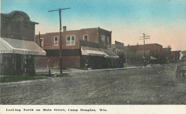 Colorized postcard of Main Street. On the left is E.J. Anthony, Barber, with a barber pole in front of the building. Other businesses can be seen, many with awnings. A man is walking on the sidewalk and the street appears to be dirt. A wagon is on the far right. Text at foot reads: "Looking North on Main Street, Camp Douglas, Wis."