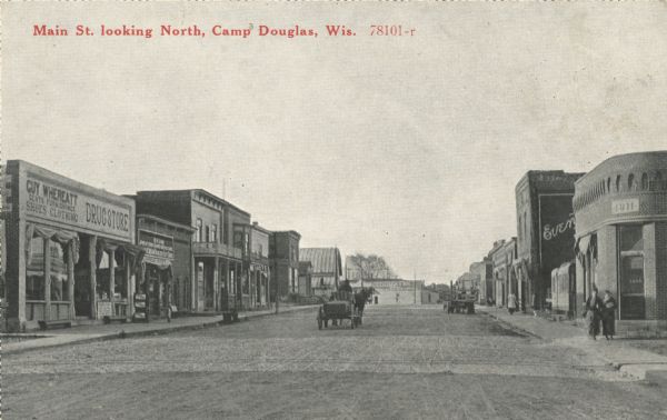 Black and white postcard of Main Street, looking north. A horse-drawn wagon is traveling north and another is parked on the right. On the left is a store, the sign reads "Guy Whereatt, Gents Furnishings, Shoes, Clothing, Drugstore." On the same side of the street is a hotel and several other businesses. On the right side are more businesses, the closest is a bank, established in 1911. The rest of the signage is not readable. Caption reads: "Main St. looking North, Camp Douglas, Wis."