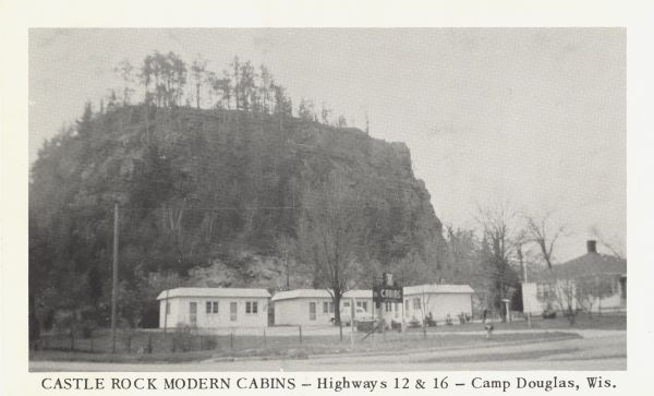 Black and white postcard of cabins in front of a bluff, perhaps Castle Rock. A larger building is visible to the right. Text on bottom reads: "Castle Rock Modern Cabins - Highways 12 & 16 - Camp Douglas, Wis."