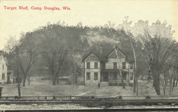 Black and white postcard of a two-story house in front of Target Bluff. In the foreground, railroad tracks and a low fence can be seen. Many trees surround the house. On the left, another house is partially visible. Text in red in upper left corner reads: "Target Bluff, Camp Douglas, Wis."