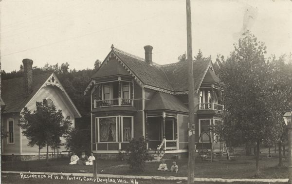 Photographic postcard of W.E. Porter's two-story house. The house has fancy trim. Three women are seated in the grass on the left, and a boy and girl are on the right. Trees surround the house. Caption reads: "Residence of W.E. Porter, Camp Douglas, Wis."