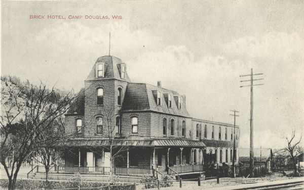Black and white postcard of the Brick Hotel (possibly the Mlakar Hotel). A large porch wraps the first floor and trees and shrubs can be seen. Text in red in upper left corner: "Brick Hotel, Camp Douglas, Wis."