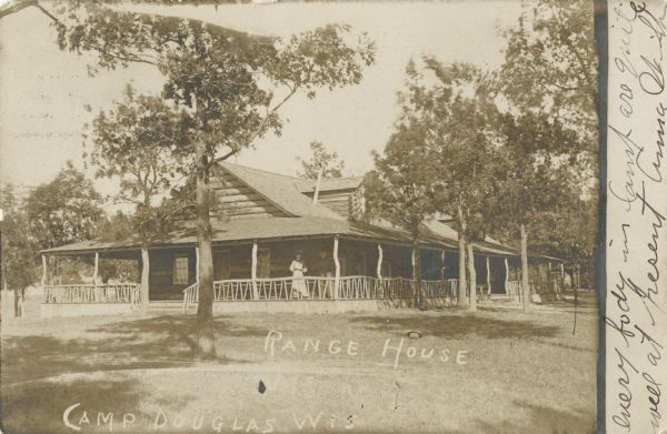 Photographic postcard of Range House, a large rustic log cabin. There is a large porch on all sides of the building with interesting roof supports and railings made of tree branches and trunks. Trees are growing in the lawn around the cabin. Two women are standing on the porch to the right of the steps, and they are wearing long dresses and hats. Caption reads: "Range House, Camp Douglas, Wis."