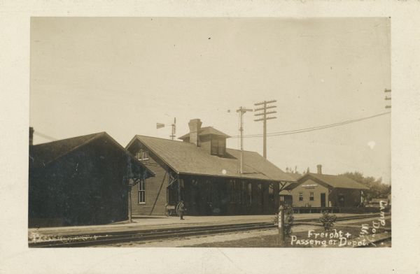 Photographic postcard of the freight and passenger depot. A young man stands on the platform, and a man can be seen entering the United States Express Company building. A windmill is behind the depot. Handwritten in lower right corner, "Freight + Passenger Depot, Camp Douglas, Wis."
