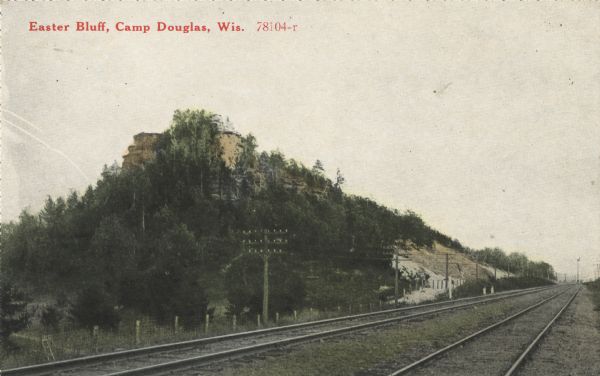 Colorized postcard of Easter Bluff, with many trees. In the foreground two sets of railroad tracks can be seen. A post and wire fence runs along the edge of the tracks and several electrical, telephone or telegraph poles are visible. Text in red in upper left corner: "Easter Bluff, Camp Douglas, Wis."