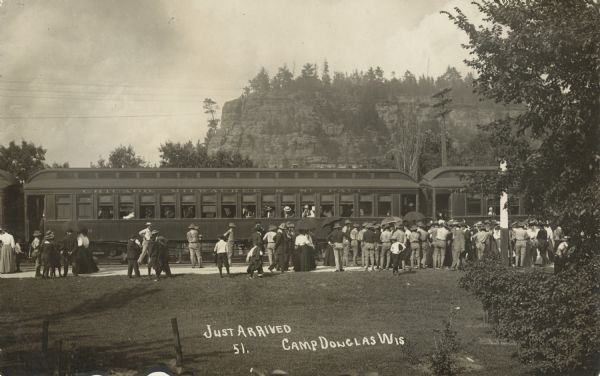 Photographic postcard of passenger train cars that have just arrived. A crowd of people are lined up waiting for the passengers to disembark, and some are holding umbrellas, presumably for protection from the sun. A tree-covered bluff rises up in the background. Handwritten below: "Just Arrived, 51. Camp Douglas, Wis."