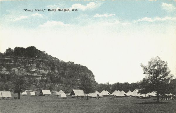 Colorized postcard of a camp scene on a lawn. In the background is a bluff. Several people can be seen standing near the tents among the trees. Caption reads: "'Camp Scene,' Camp Douglas, Wis."