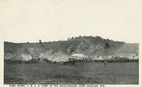 Black and white postcard of Camp Douglas with the Y.M.C.A. Camp in the background. Many tents are pitched and soldiers are lined up on the left. An automobile is parked in the center. Bluffs are in the background. Caption reads: "Camp Scene, Y.M.C.A. Camp in the back-ground, Camp Douglas, Wis."