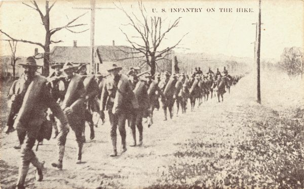 Postcard printed in brown ink of a line of soldiers in uniform hiking in a column. They have bedrolls over their shoulders. Towards the rear are soldiers on horseback. Trees and buildings are in the background. Text at the top reads: "U.S. Infantry On The Hike."