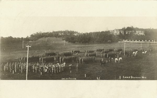Photographic postcard of elevated view of soldiers during a dress parade. Some soldiers are on horseback, but most are on foot. There is a musical band in the lower left corner. In the background are bluffs. Handwritten at foot: "Dress Parade, 21, Camp Douglas, Wis."