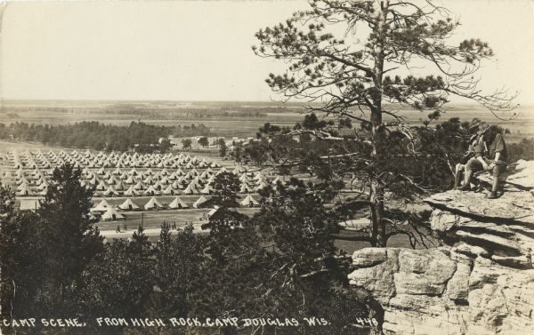 Photographic postcard of an elevated view of a camp scene. Tents are pitched in rows with roads in between. Soldiers are walking along the roads.  Two soldiers are sitting on the top of the rock formation (possibly High Rock) on the right, and one of them is holding a piece of radio equipment or a camera. Handwritten at the foot: "Camp Scene, From High Rock, Camp Douglas, Wis."