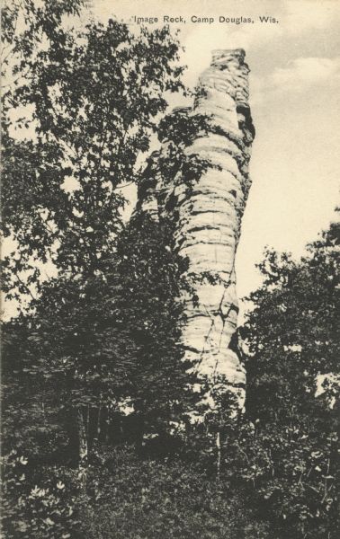 Black and white postcard of Image Rock, seen through a stand of trees. Text at top reads: "Image Rock, Camp Douglas, Wis."
