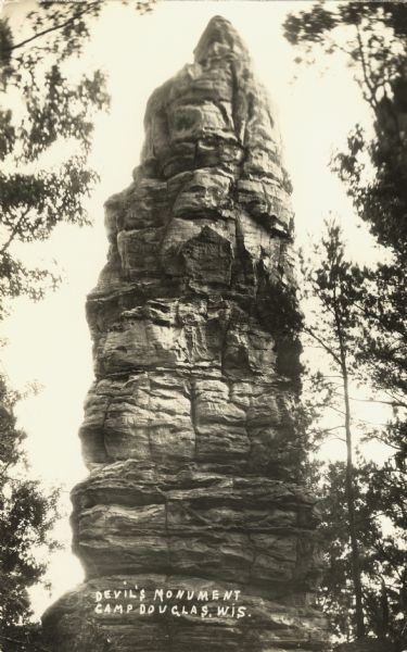 Photographic postcard view of Devil's Monument. Trees are on both sides. Text handwritten at foot: "Devil's Monument, Camp Douglas, Wis."