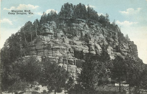 Colorized postcard of Magazine Bluff. Trees are on top of the bluff and also below. A stone wall is among the trees in the lower right corner. Text in upper left reads: "Magazine Bluff, Camp Douglas, Wis."