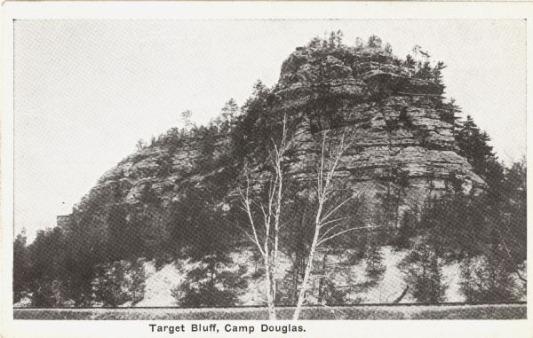 Black and white postcard of Target Bluff. Trees are on top of the bluff and also below. Railroad tracks are at the base. Text at foot reads: "Target Bluff, Camp Douglas."