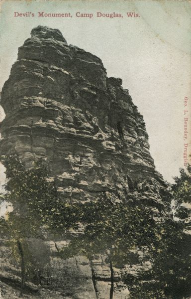 Colorized postcard of Devil's Monument with trees surrounding it. Text at top reads: "Devil's Monument, Camp Douglas, Wis."
