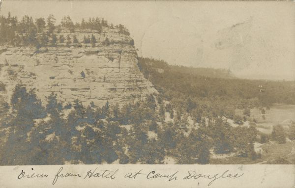 Photographic postcard view of Long Bluff. Trees are on top of the bluff and also below. More bluffs and trees are in the background. Handwritten below: "View from Hotel at Camp Douglas."