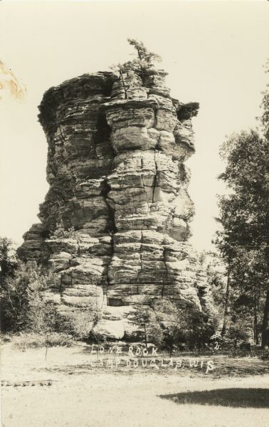 Photographic postcard of Lone Rock. Trees are on top and also around the base. Handwritten at foot: "Lone Rock. Camp Douglas, Wis."