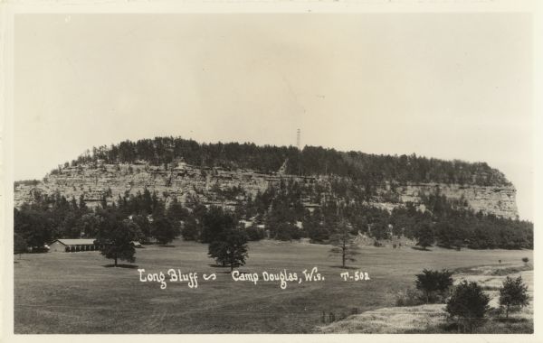 Photographic postcard elevated view over field of Long Bluff. Trees and a watchtower are on top of the bluff. In the lower left corner is a long building with open sides. Text at foot reads: "Long Bluff - Camp Douglas, Wis. T-502."
