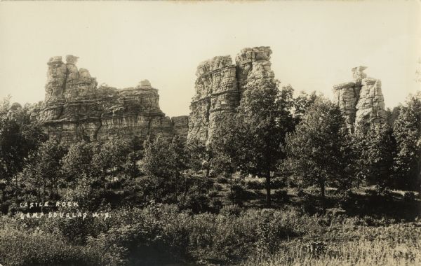 Photographic postcard of Castle Rock. Elevated view over trees and shrubs in the foreground. Text in lower right corner reads: "Castle Rock, Camp Douglas, Wis."