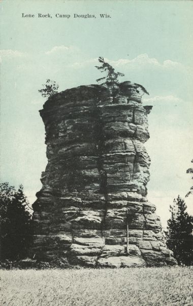 Colorized postcard looking up towards Lone Rock. Trees are on top of the formation. Caption reads: "Lone Rock, Camp Douglas, Wis."