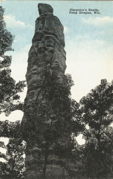 Colorized postcard of Cleopatra's Needle with trees all around the base. Text at top reads: "Cleopatra's Needle, Camp Douglas, Wis."