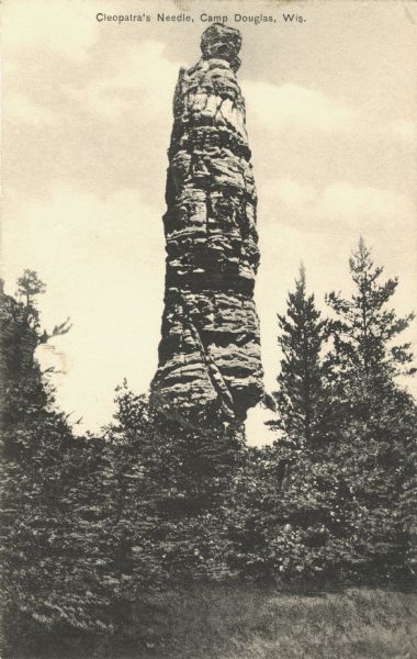 Black and white postcard of Cleopatra's Needle with trees all around the base. Text at top reads: "Cleopatra's Needle, Camp Douglas, Wis."