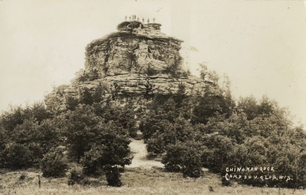 Photographic postcard of Chinaman Rock. Trees surround the base and eight human figures can be seen standing on the summit, after hiking or climbing to the top. Text in lower right corner reads: "Chinaman Rock, Camp Douglas, Wis."
