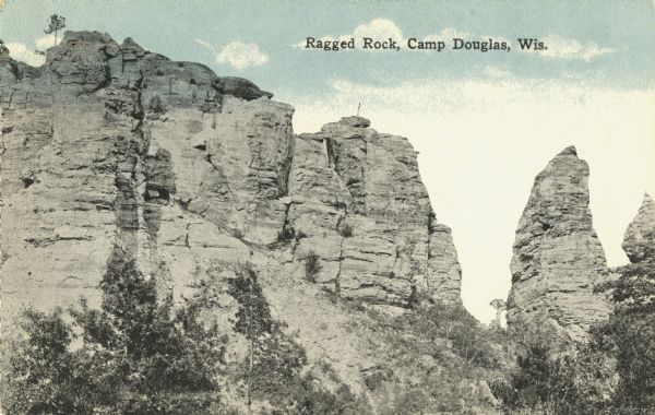 Colorized postcard of Ragged Rock in Mill Bluff State Park. It is an 80 foot tall former bluff whose protective cap was worn away and is eroding in a conical mound. Trees are at the base. Text at top reads: "Ragged Rock, Camp Douglas, Wis."