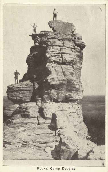Black and white postcard of a rock spire with four men posing in various places. Trees are in the background. Text at foot reads: "Rocks, Camp Douglas."