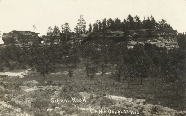 Photographic postcard of Signal Rock. Trees are in the foreground and on the the hill. Text below reads: "Signal Rock, Camp Douglas, Wis."