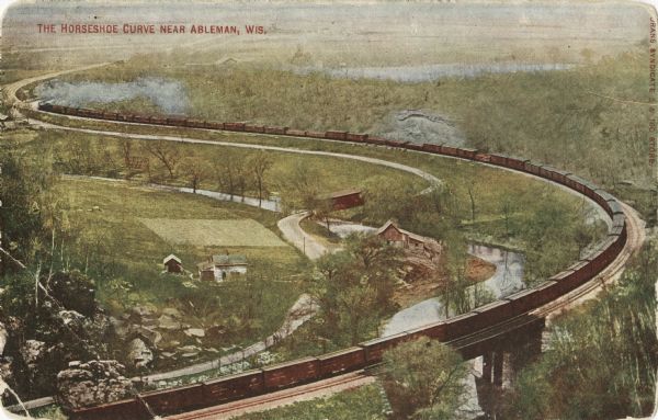 Colorized postcard of the horseshoe curve of railroad tracks. Elevated view of the railroad tracks with train snaking past a farmhouse and a bridge suspended over a river. There is a covered bridge over the river, and a lake is in the background.