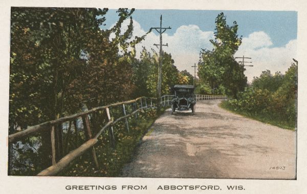 Colorized postcard of a car coming down a road. There is a fence on the left near trees, and there are telephone poles in the background. Caption reads: "Greetings From Abbotsford, Wis."