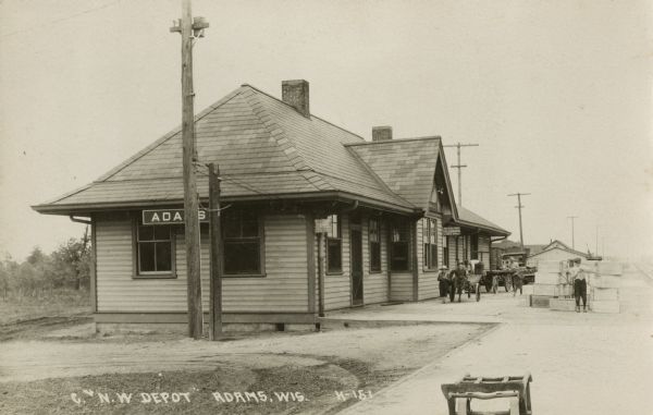Photographic postcard of the Chicago and North Western Depot. A group of children are in front of the depot near a cart, and two men work nearby.