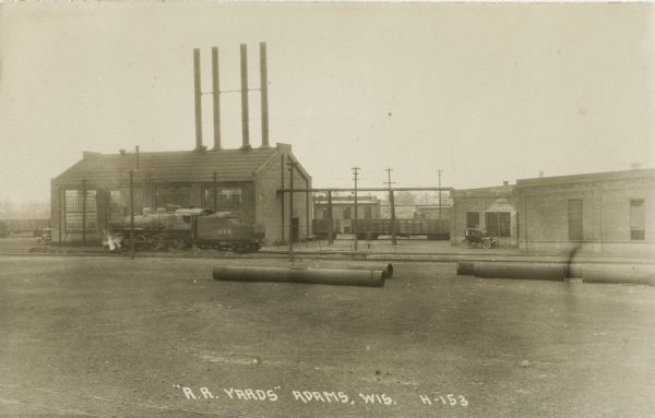 Photographic postcard of the railroad yard. Across an empty yard where a few large pipes lay, a locomotive with smoking stack and cars is on railroad tracks in front of brick buildings with tall smokestacks. One car is labeled Chicago and North Western, the other says Chicago, Saint Paul, Minneapolis and Omaha.