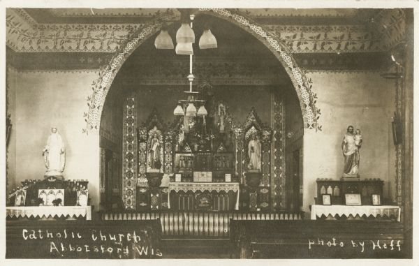 Interior of a Catholic church. View of front of the church with the altar and religious icons. There are ornate designs near the ceiling and the arch to the altar. Chandeliers hang from the ceiling, and there are pews on either side of the main aisle. Caption reads: "Catholic Church, Abbotsford, Wisconsin."