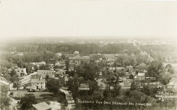 Photographic postcard of the cities of Adams and Friendship. Elevated view of the downtown area, including tree-lined streets, city businesses and homes.