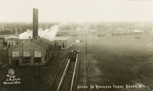 Photographic postcard of the railroad yards. Elevated view of railroad tracks and switch-yard, with steam rising behind a brick building on the left. Railroad cars and rural farmhouses can be seen in the background.