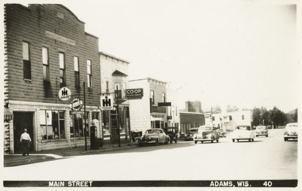Photographic postcard of Main Street. Businesses line the street, including the Adams Auto Company Dodge-Plymouth, McBride Equipment Company, and McCormick International Harvester Company. There is also a food co-op and a Hudson standard service station.