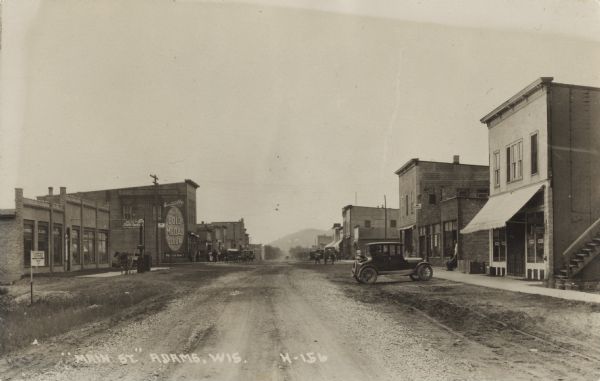 Photographic postcard of Main Street. People, automobiles, and horse-drawn vehicles line the dirt-covered street and sidewalks. A Gold Medal Flour billboard and a Ford automobile sign are on the side of a building on the left. A camp grounds sign is in the left foreground. In the far background is a hill or bluff.