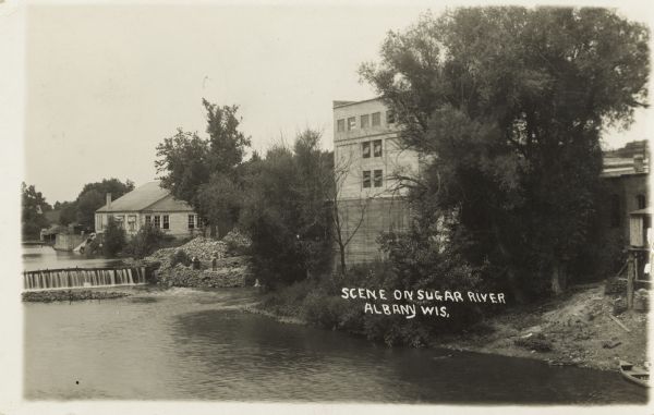 Photographic postcard of the Sugar River. Elevated view of buildings and trees lining the shoreline. Two men can be seen standing next to the dam among a pile of bricks. Caption reads: "Scene on Sugar River, Albany, Wisconsin."