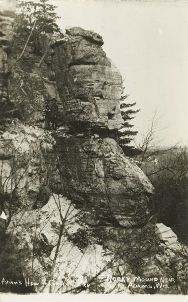 Photographic postcard of a geological rock formation. Side view of the snow-covered bluff and rocks surrounded by trees. Two children stand on a ledge next to the rock outcropping. The image is of the south side of the gap of Rattlesnake Mound, just south of Adams