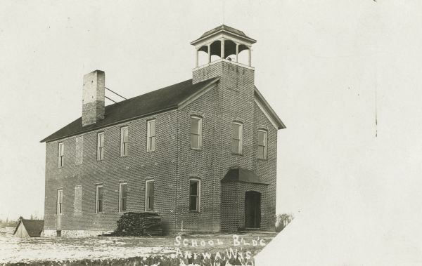 Photographic postcard of a two-floor school building with a covered front entrance underneath a bell tower. Snow is on the ground. Caption reads: "School B'LD'G, Aniwa, Wis."