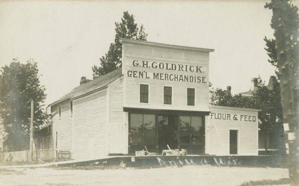 Photographic postcard of the entrance to the G.H. Goldrick General Merchandise store. Trees and a flier-covered telephone pole can also be seen. Signs on the front of the building reads, "G.H. Goldrick, Gen'l Merchandise" and "Four & Feed." Caption reads: "Aniwa, Wis."